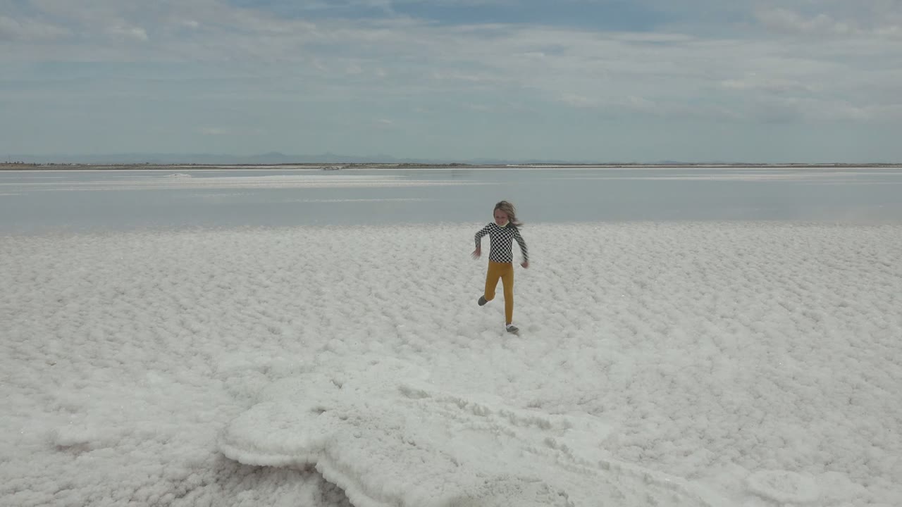 A child runs across the white salt flat in Guerrero Negro, Baja California Sur, with shallow water and a cloudy sky behind them.