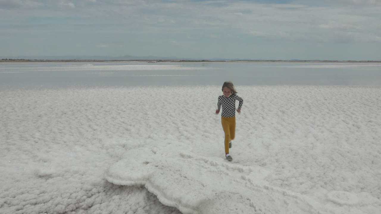 The child moves closer across the textured white salt crust in Guerrero Negro, with a flat waterline and pale clouds in the distance.
