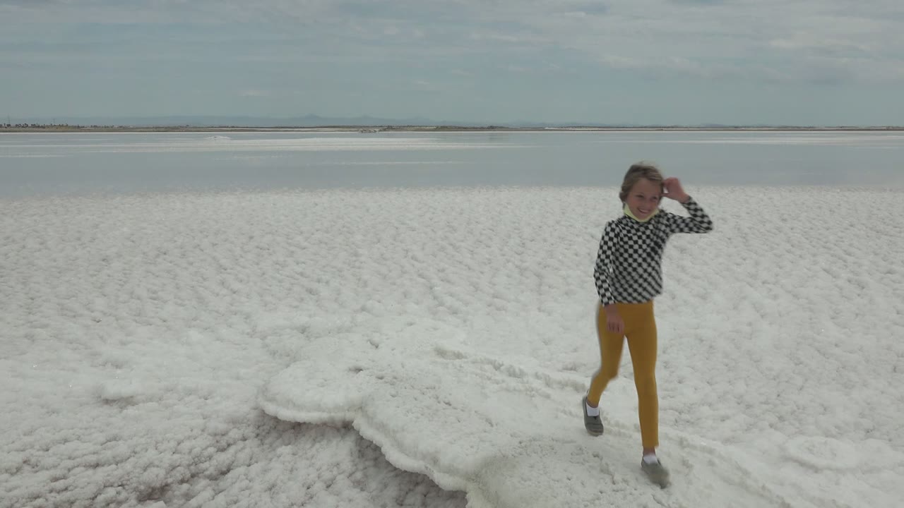 The child reaches the right side of the Guerrero Negro salt-flat view, with patterned white ground, calm water, and a broad cloudy sky behind them.