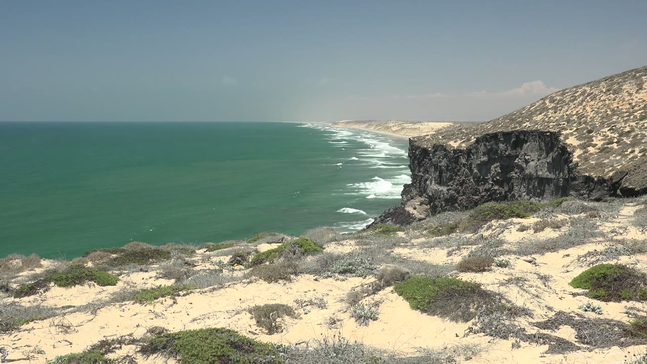 San Quintin BC Mexico coastline with turquoise ocean, white surf, sandy scrub foreground, a dark cliff at right, and no visible people at the opening moment.
