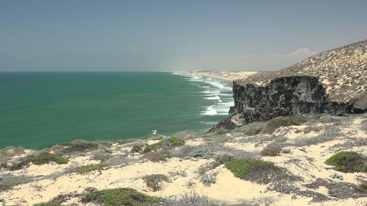 Turquoise water meets a sandy and rocky San Quintin BC Mexico shoreline with scrub in the foreground, a dark cliff to the right, and no visible people.