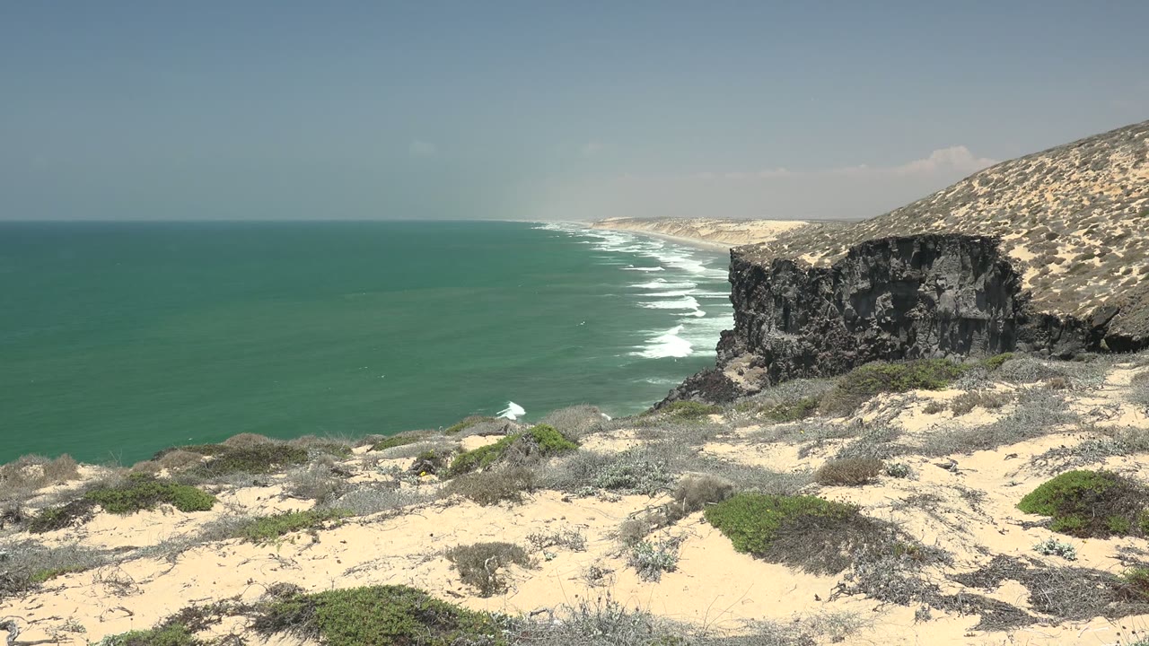 White surf lines run along a sandy coast below a dark rock cliff.
