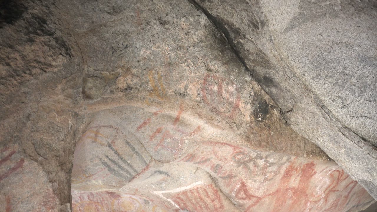Faded red symbols and black marks appear beneath a gray stone overhang on the rock art surface in Santa Rosalia, with no visible people.