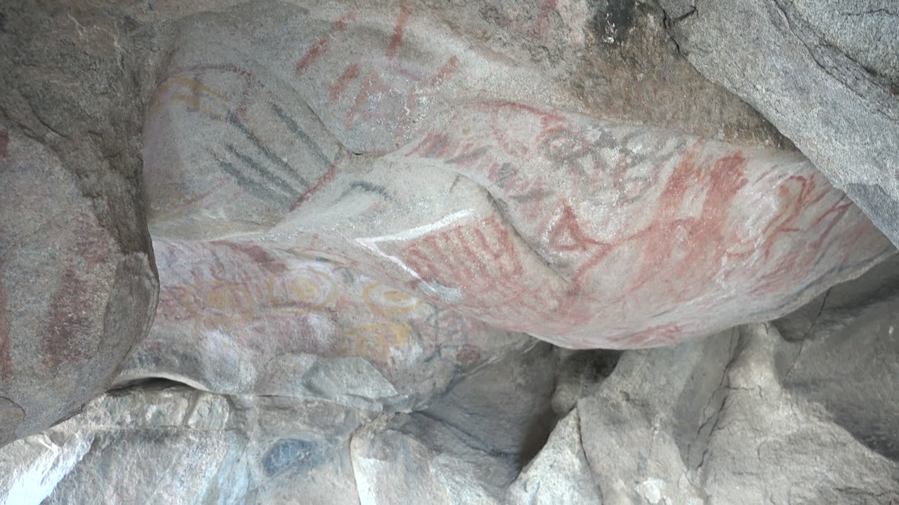 Opening frame of ancient rock paintings on a gray cave ceiling in Santa Rosalia, Baja California Sur, with red pigment and darker line marks visible and no visible people.