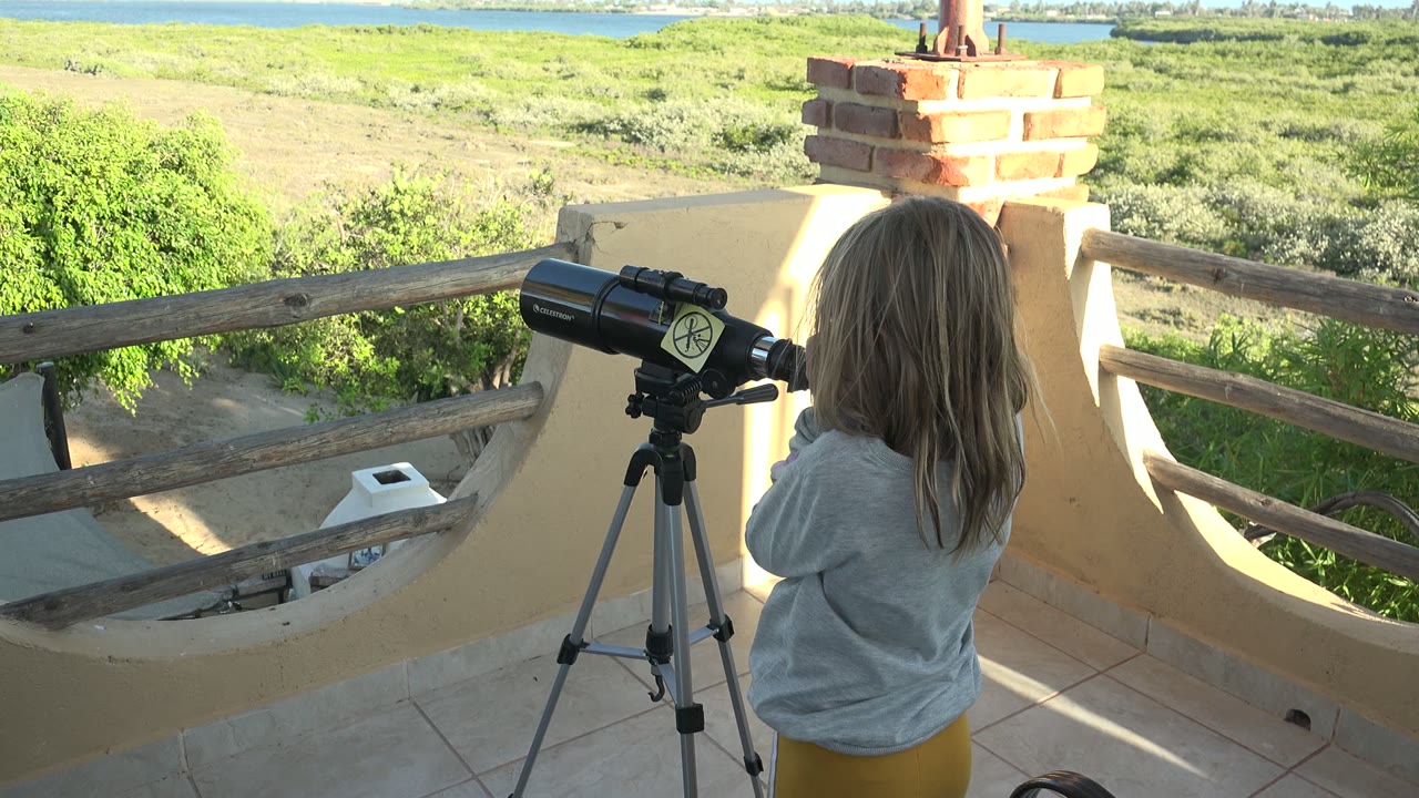 The child stands close to the black telescope near a brick column on the San Carlos platform, with sunlit tile, wood rail, shrubs, sandy terrain, and water visible behind.