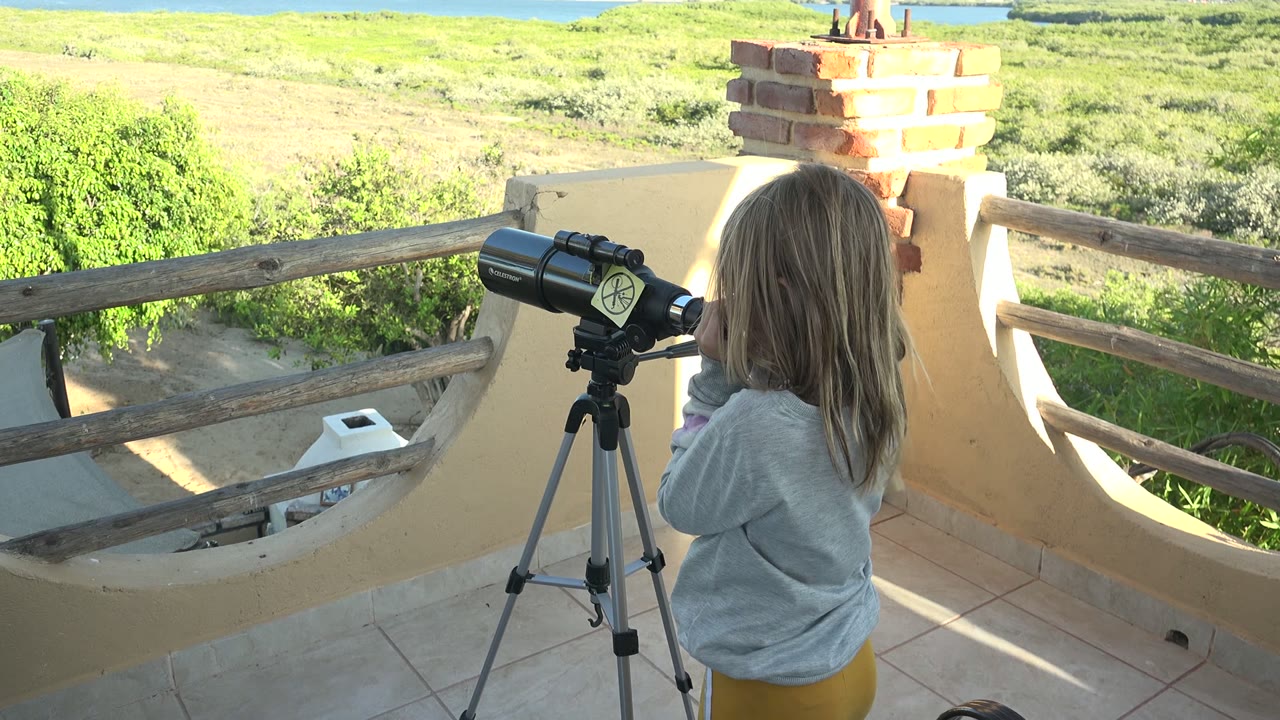 At 0:00 in San Carlos, Baja California Sur, a young girl stands at a tripod-mounted telescope on a balcony with bright green vegetation and distant water beyond the railing.