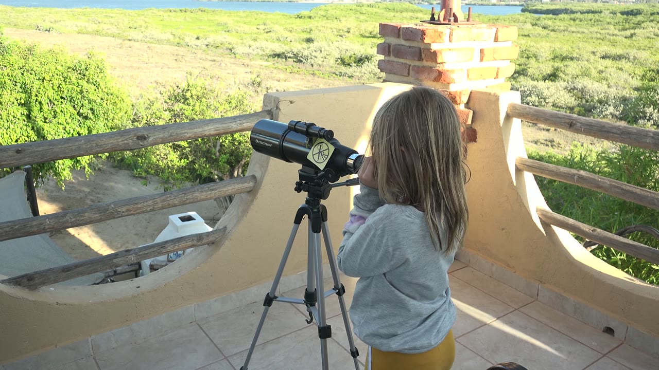 Child at a black telescope on a sunlit balcony with a wooden railing.