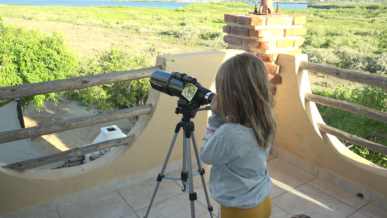 At 0:02 in San Carlos, Baja California Sur, the girl in yellow pants looks into the telescope near the wooden rail, with open green land and a strip of water in the distance.