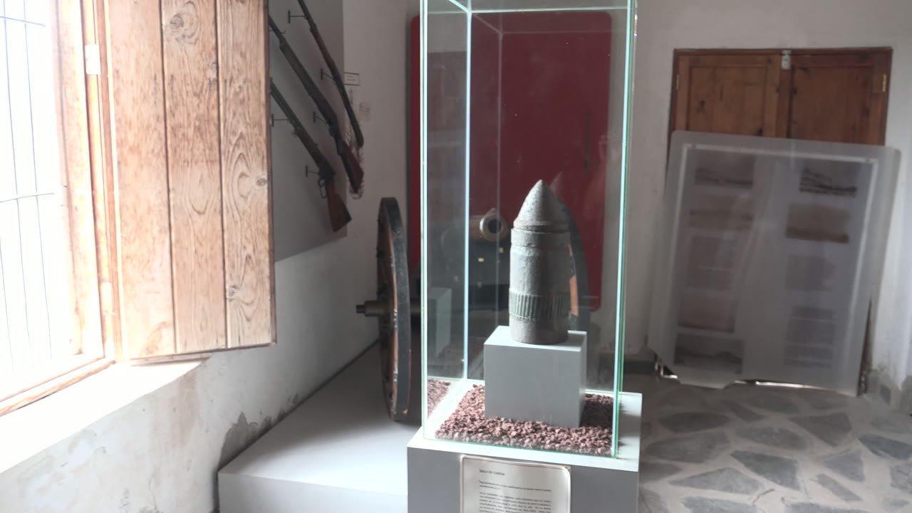 A dark artifact in a glass display case stands on a white pedestal inside a Mulege, Baja California Sur museum room, with open wood shutters, a red door, and no visible people.
