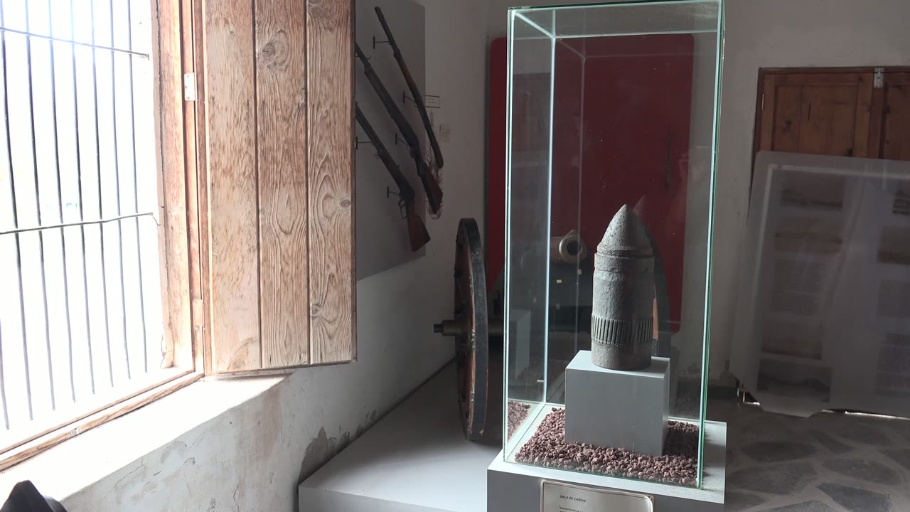 A protected artifact on a pedestal is seen through the glass case in Mulege, Baja California Sur, with bright vertical window bars, wood shutters, and no visible people.