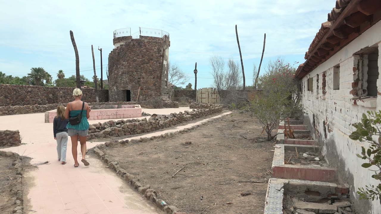 In Mulege, Baja California Sur, a woman and child walk away on a pale pink path toward a rough stone tower beside dry ground and an old white wall.