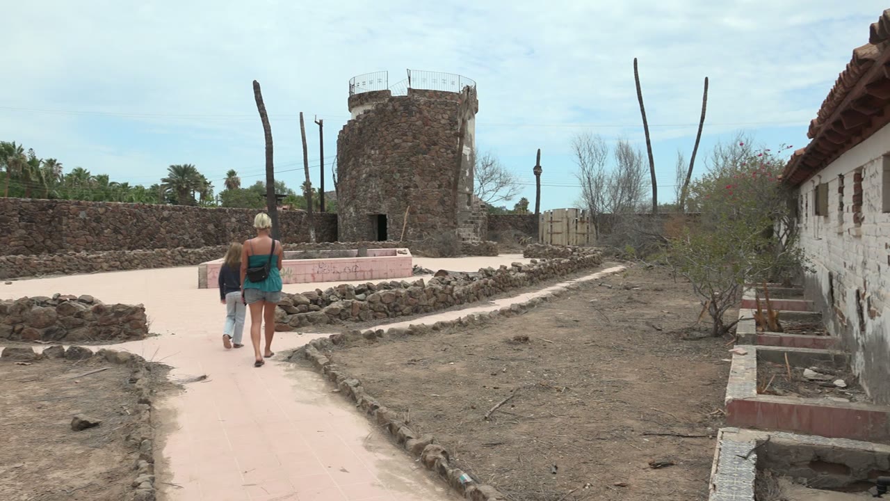 The walkers are slightly farther from camera on the curving path, with the white wall on the right edge and the stone tower set beyond the low rock edging.