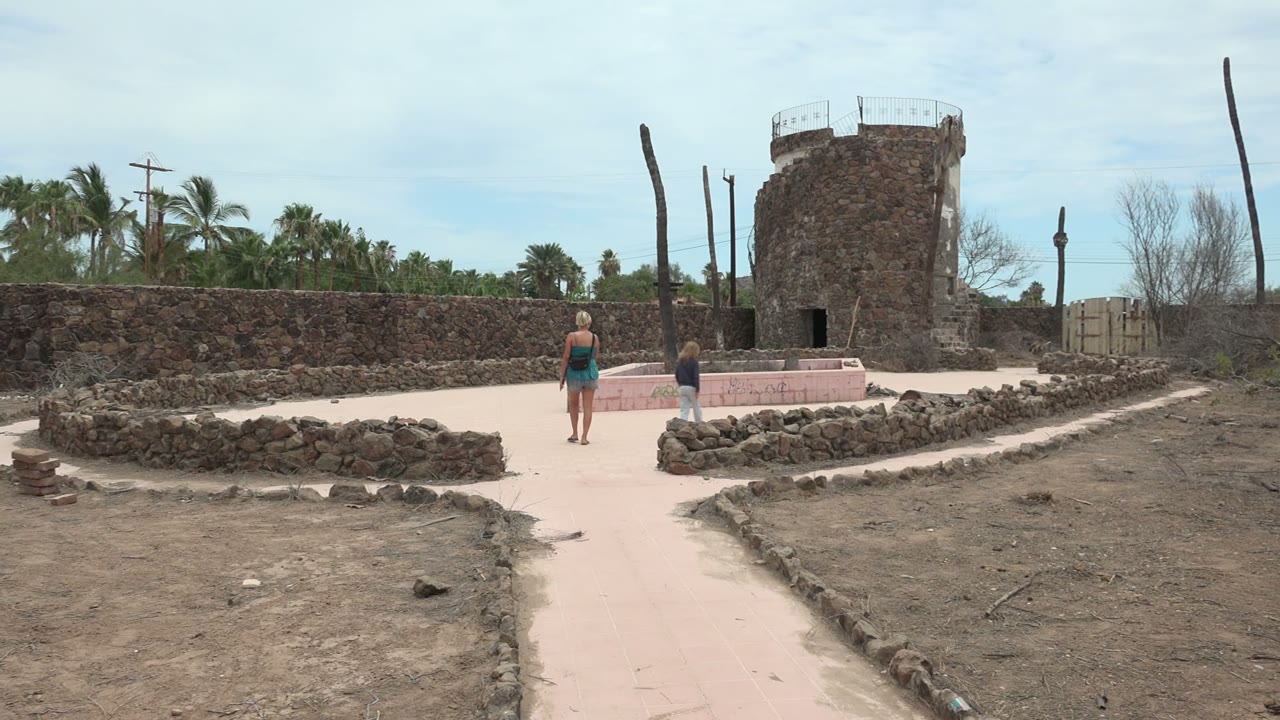 At the opening moment in Mulege, Baja California Sur, two people walk away from the camera along a pale sandy path toward a round stone tower and low stone walls.