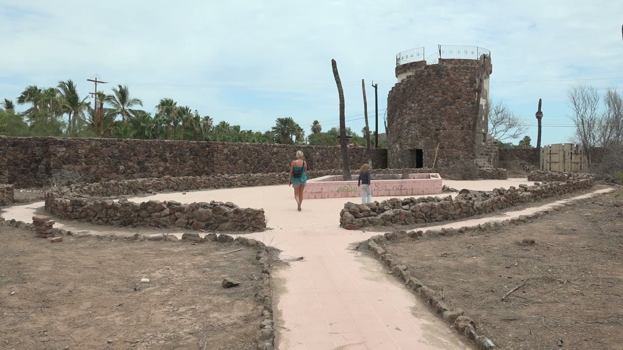 Two people farther along the sandy path near a stone tower and low rock borders.