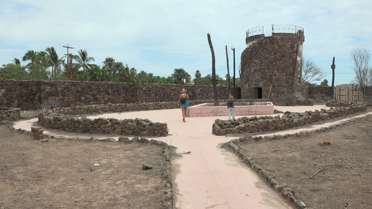 At 0:02, the walkers continue toward the stone tower in Mulege, with dark stone walls stretching across the frame and palm trees visible beyond the courtyard.