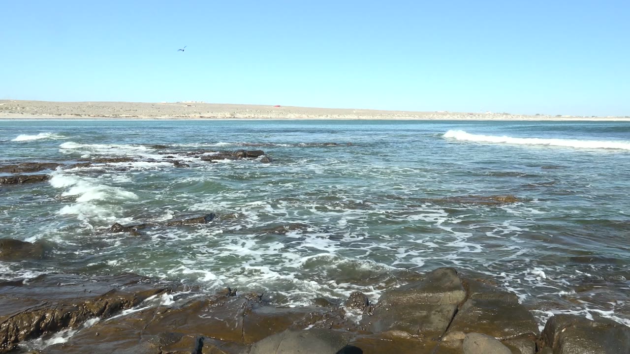 At 0:00 in La Bocana, Baja California Sur, ocean foam spreads over dark foreground rocks while a low distant shoreline sits beneath a clear blue sky, with no visible people.