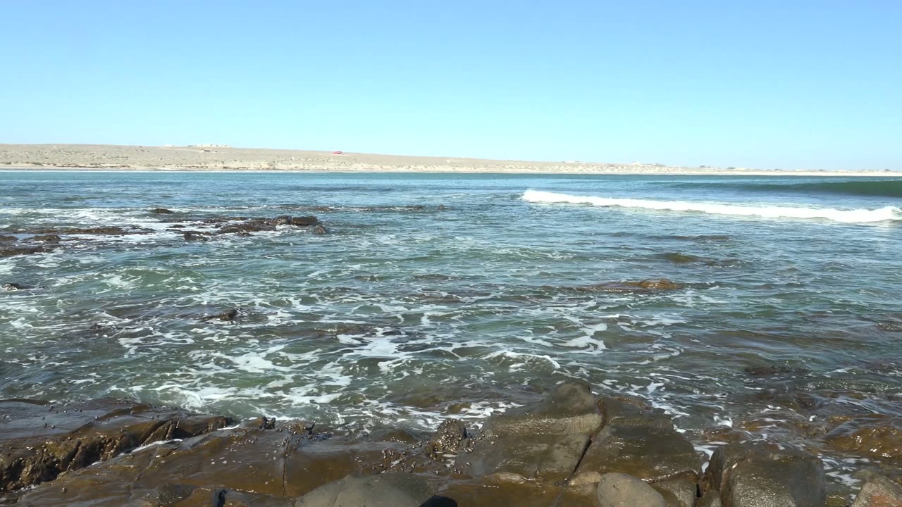 At 0:01 in La Bocana, Baja California Sur, a white wave line breaks farther offshore while wet rocks remain visible in the foreground and no people are visible.