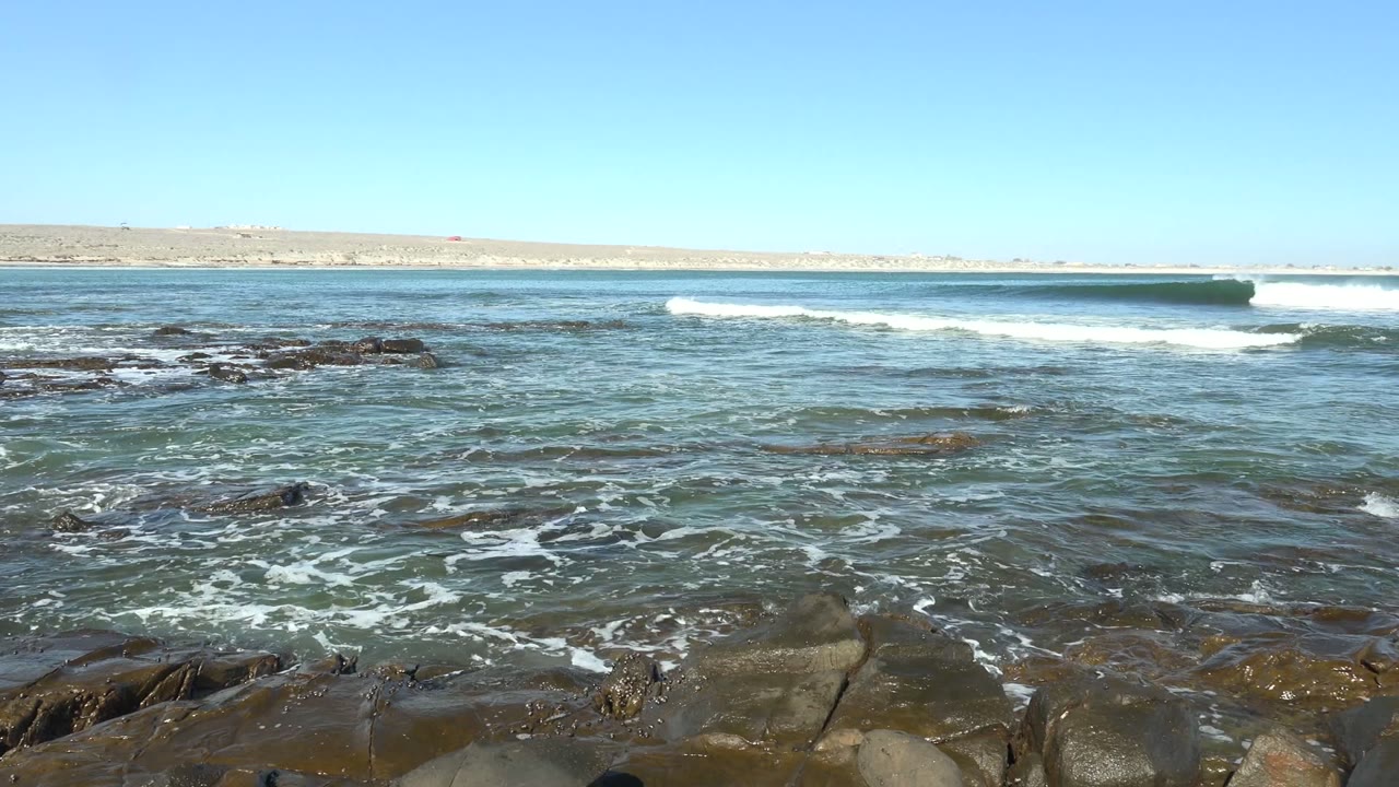 At 0:02 in La Bocana, Baja California Sur, a low swell crosses the midground as broken foam patterns spread around exposed rocks, with no visible people.