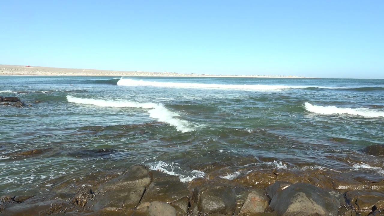 At 0:00 in La Bocana, Baja California Sur, white waves break across dark foreground rocks with a distant landmass on the horizon and no visible people.