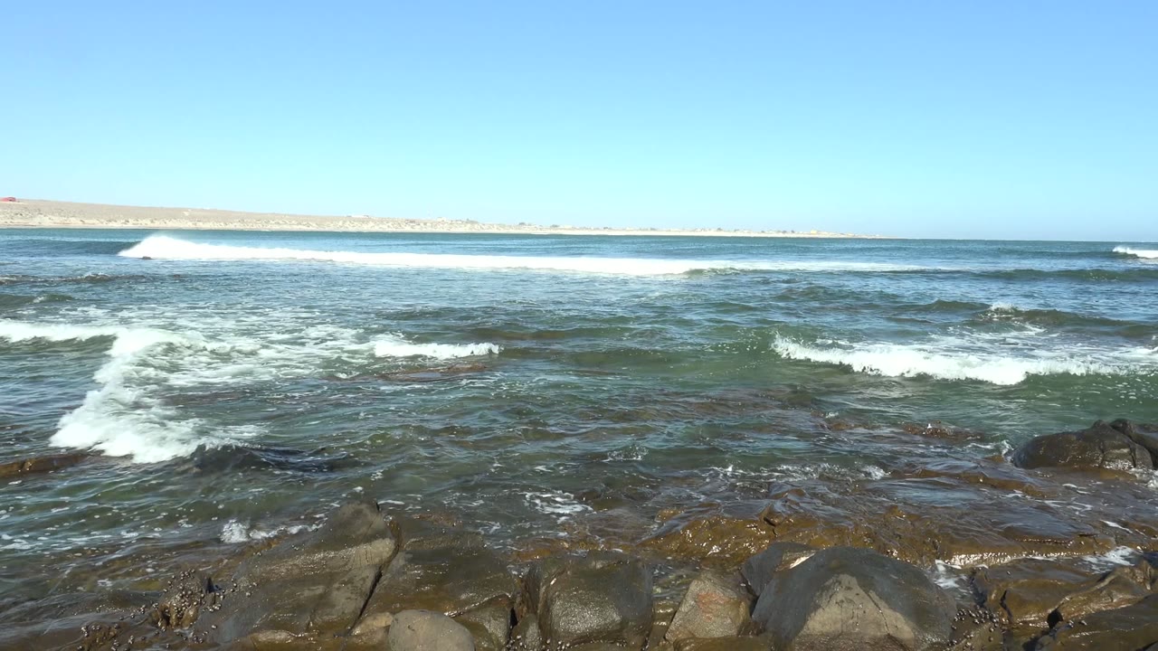 At 0:02 in La Bocana, Baja California Sur, a swell breaks left of center while smaller whitewater lines cross the rocky foreground, with no visible people in the frame.