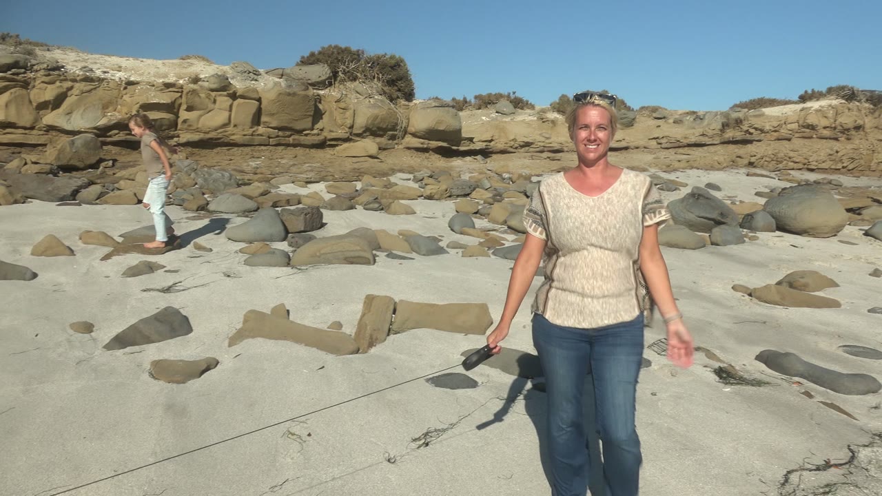 At La Bocana in Mexico, a smiling woman stands close to the camera on pale sand with scattered rocks while a man walks behind her along the rocky beach.