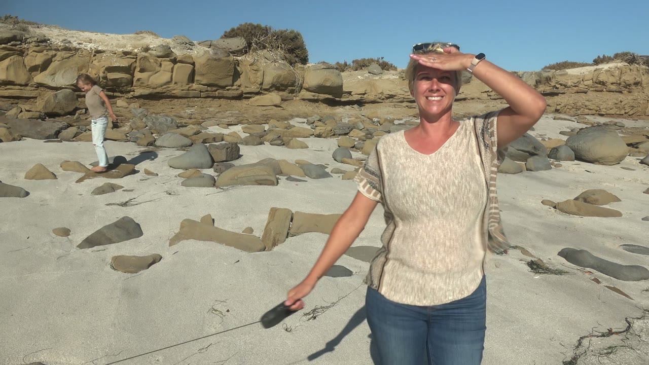 Woman saluting in bright sun as a man walks across the rocky sand behind her.