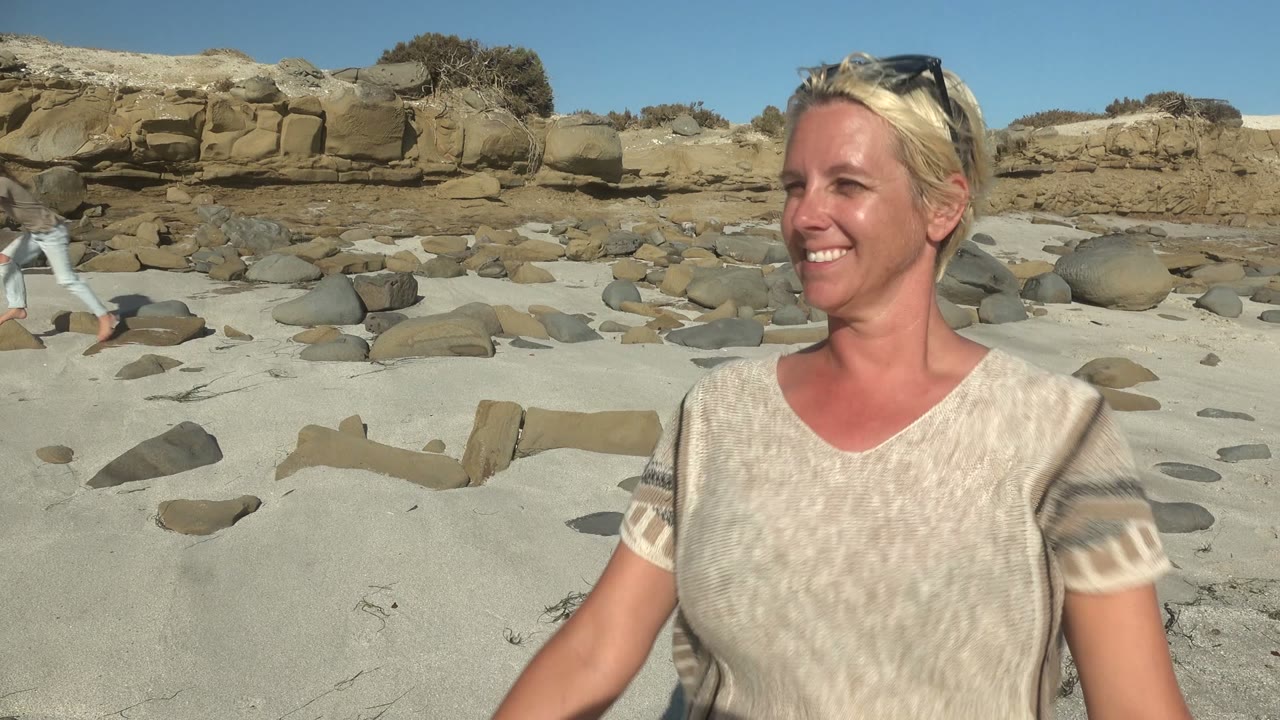 At La Bocana in Mexico, the woman's face and upper body fill the right side of the frame while the sandy, rock-strewn beach and low coastal ledge remain visible behind her.
