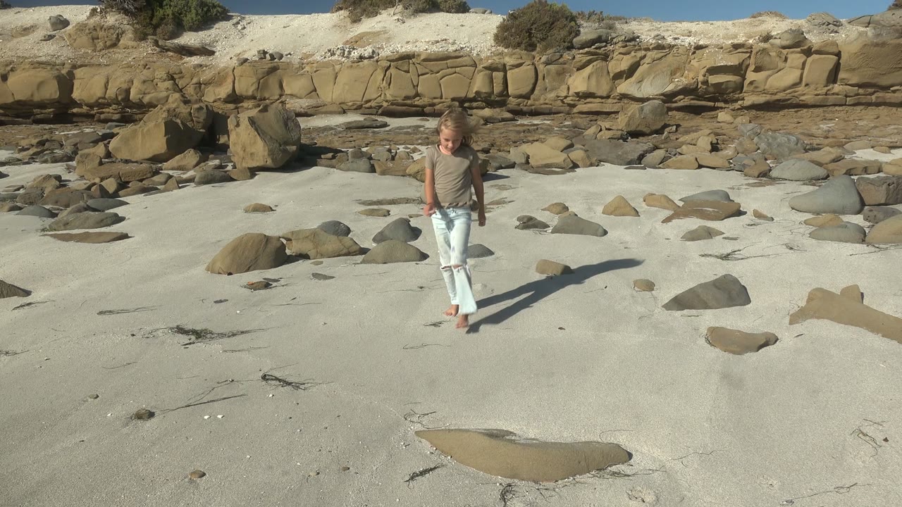 A child runs barefoot toward the camera on a sandy La Bocana beach, with scattered rocks, tan dunes, sparse vegetation, and a long shadow visible in bright sun.