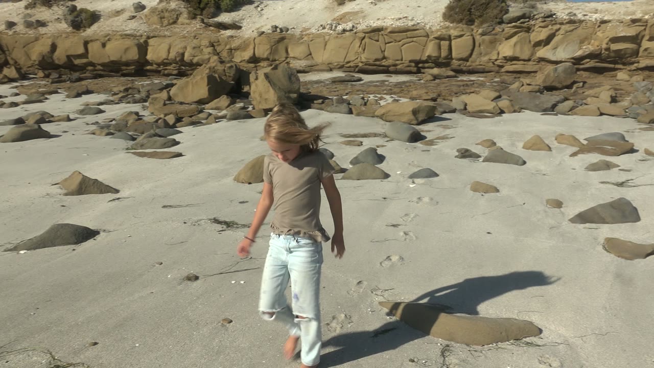 The child appears near the right side of the La Bocana beach frame, looking down as footprints, scattered rocks, pale sand, and the eroded dune wall fill the scene.