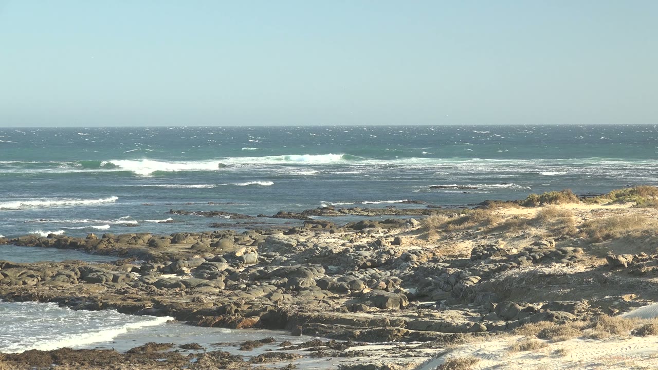 Wide view of waves breaking across a rocky La Bocana shoreline with dry grass in the middle ground, blue water beyond, and no visible people.