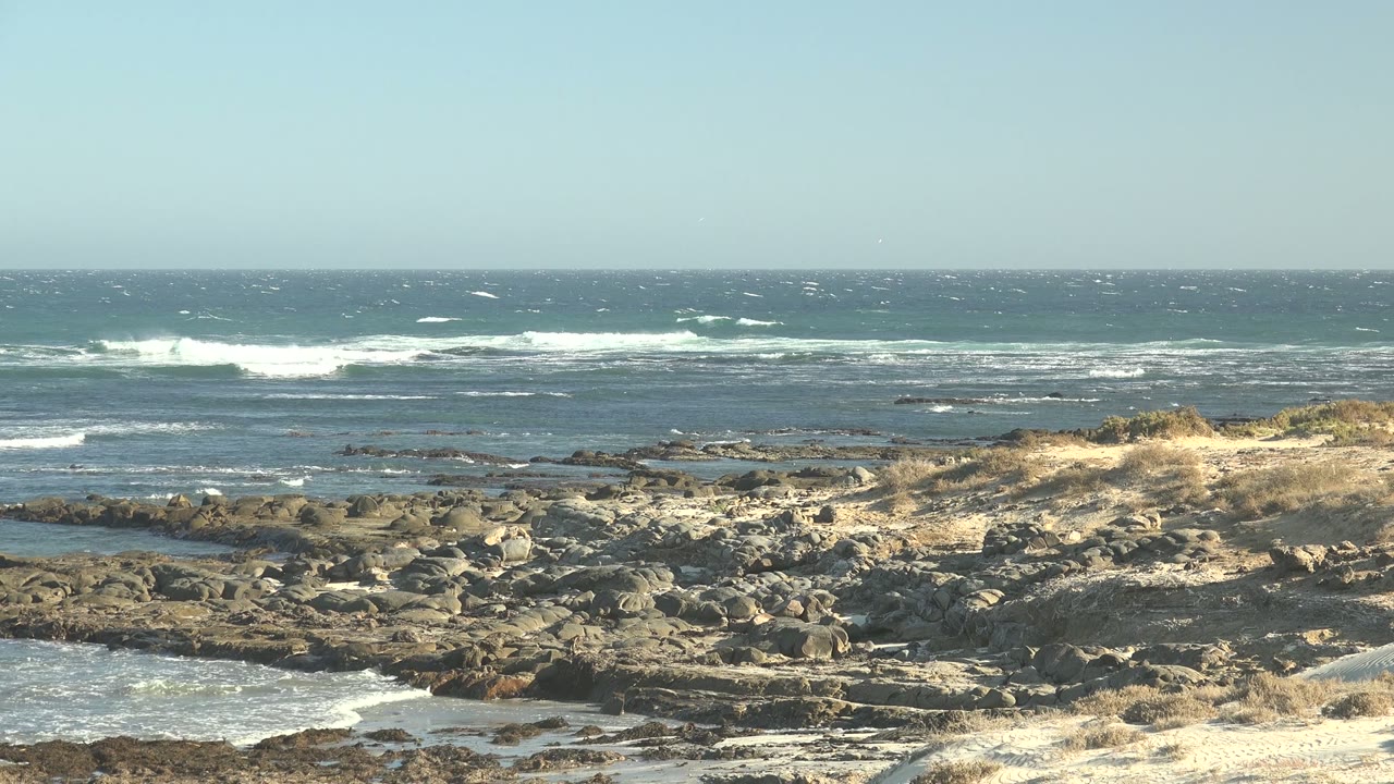 A wide La Bocana coastal frame shows foamy surf behind dark rocks, sunlit dry grass near the center, and an empty shoreline with no visible people.