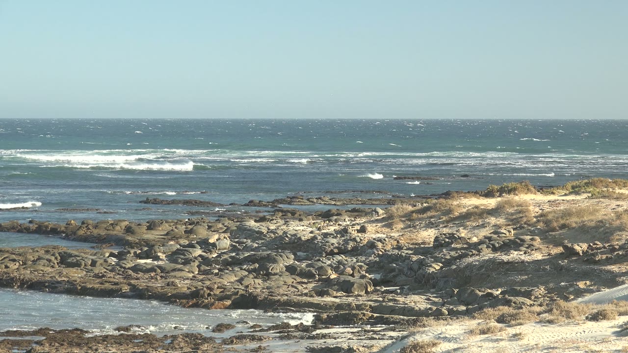 Opening frame of La Bocana, Baja California Sur, with white surf breaking beyond a rocky shoreline, sandy foreground, dry grass, and no visible people.
