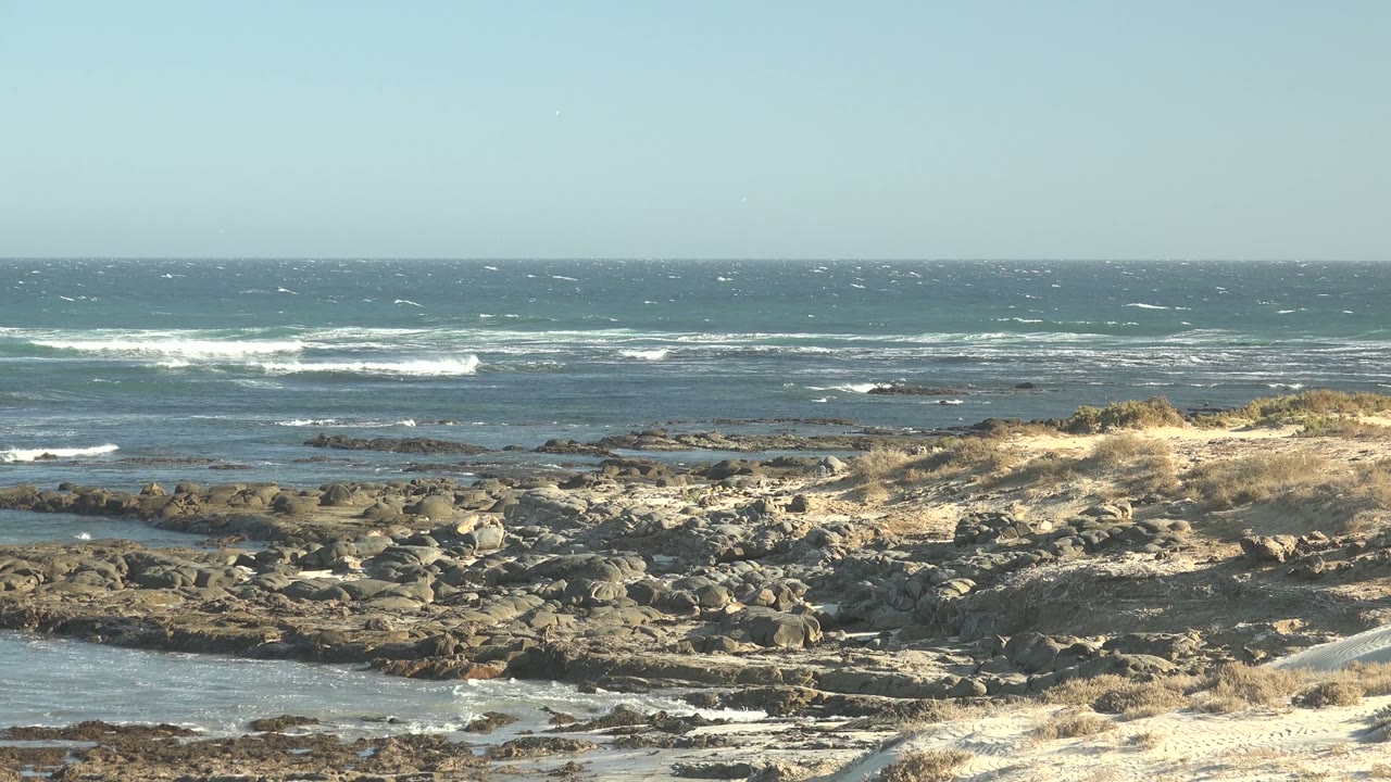 Ocean surf breaks farther offshore behind rocks, sand, and dry grass.