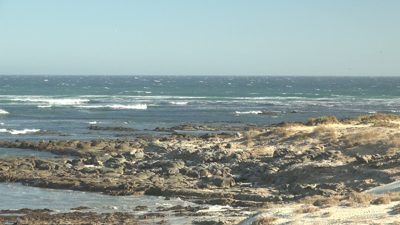 Third frame holds the La Bocana coastline as choppy blue water and white wave crests sit behind sunlit sand, dry grass, scattered rocks, and no visible people.
