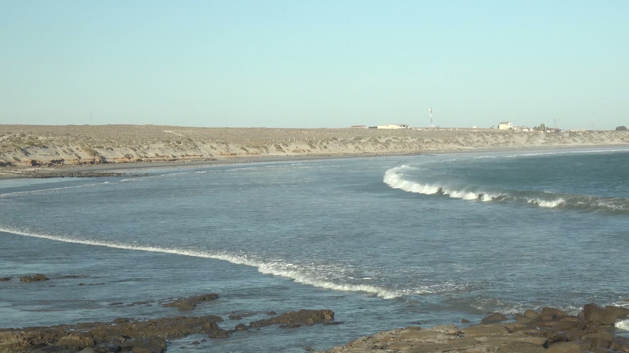 Wide view of ocean waves approaching a rocky shoreline in La Bocana, Baja California Sur, with dry land and pale sky behind, no visible people.