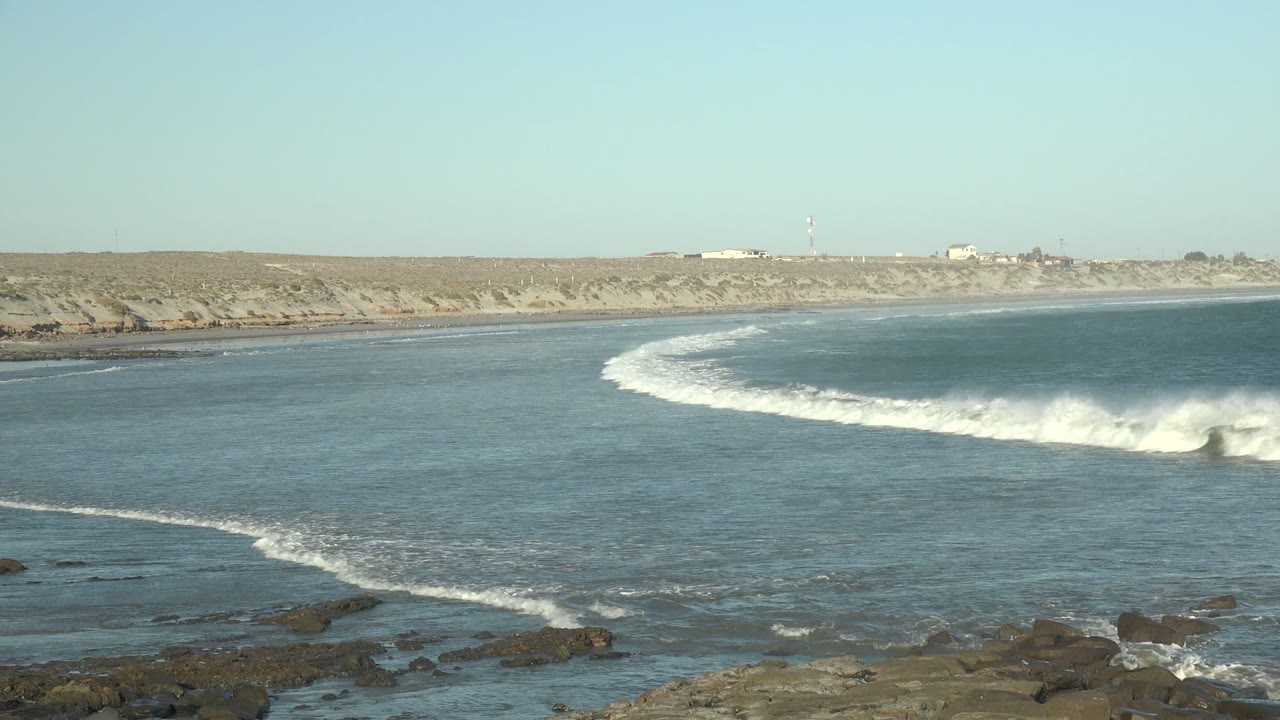 White surf breaks more strongly along the right side of the La Bocana shoreline while rocks remain visible in the lower foreground, no visible people.