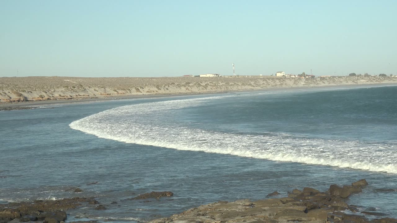 At 0:00 in La Bocana, Baja California Sur, white foam arcs across blue-green water toward dark shoreline rocks, with sandy dunes beyond and no visible people.