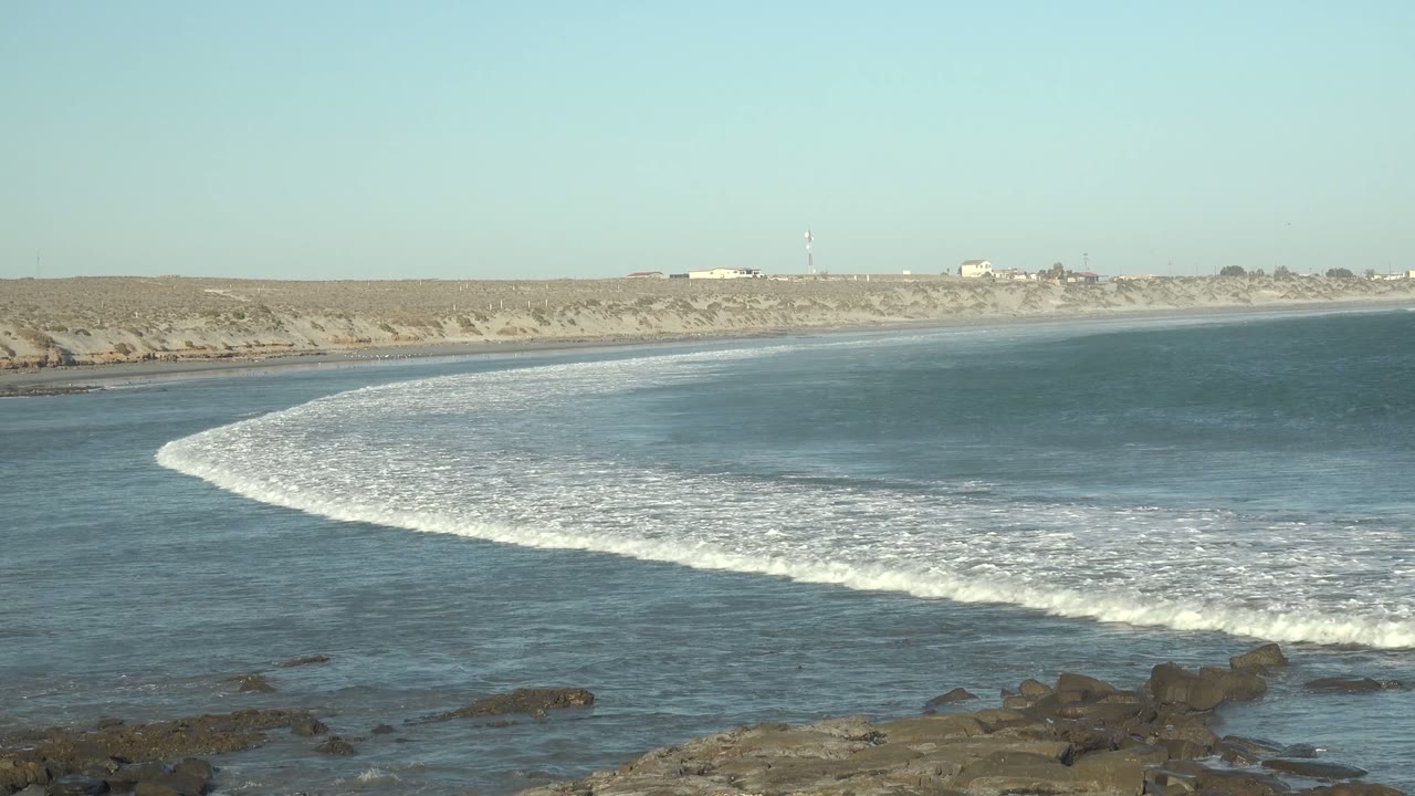 White surf line spreads across coastal water near dark rocks.
