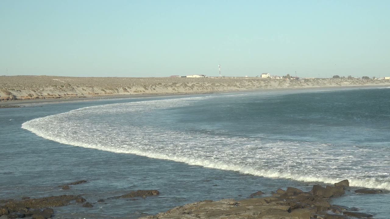 At 0:02 in La Bocana, Baja California Sur, the breaking wave spreads white foam through the midground with exposed rocks below, sandy dunes behind, and no visible people.
