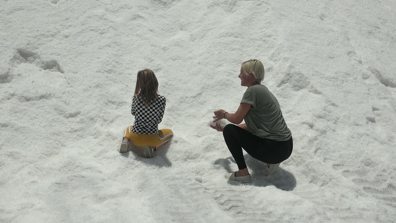 At 0:00 in Guerrero Negro, Mexico, a child sits facing away on a yellow sled while an adult crouches to the right on the bright snowy slope.