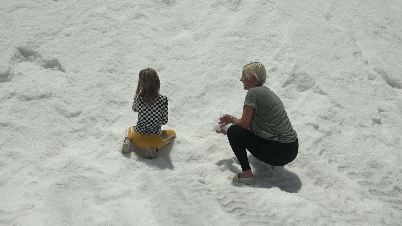 Child seated on yellow sled as an adult crouches nearby in snow.