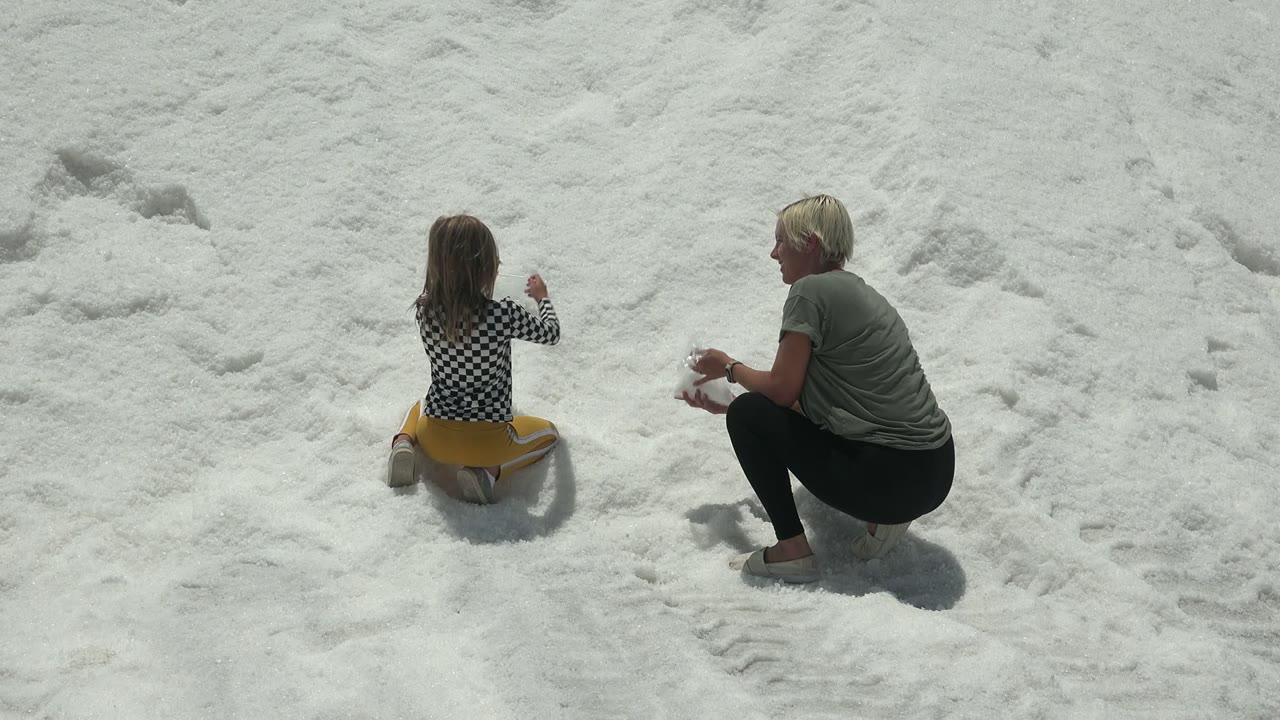 At 0:02 in Guerrero Negro, Mexico, the child lifts an arm while seated on the yellow sled and the adult watches from a crouched position on the snowy slope.