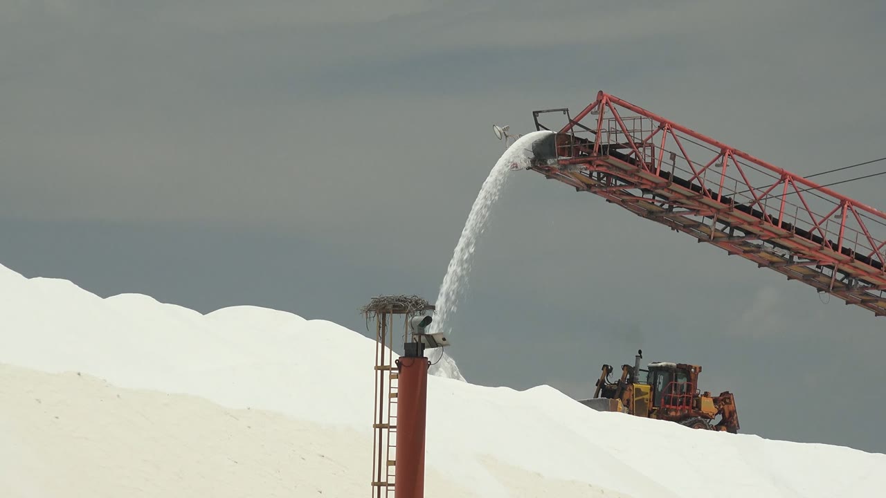Conveyor pours salt over a white mound with machinery below.