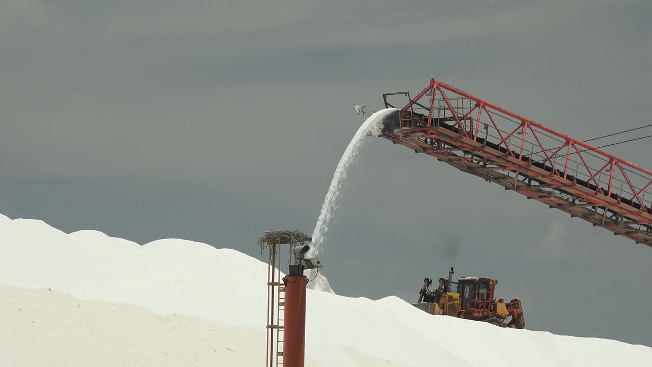 The red conveyor reaches across the upper frame as white salt pours down in Guerrero Negro, Baja California Sur, while a yellow vehicle and distant workers sit low behind the pile.