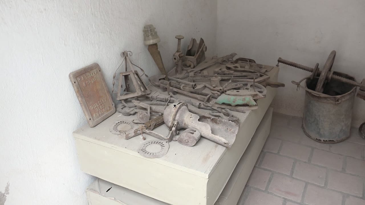 Dusty tools and metal pieces rest on a pale cabinet in a Mulege workshop corner, with a bucket at right, white walls, brick floor, and no visible people.
