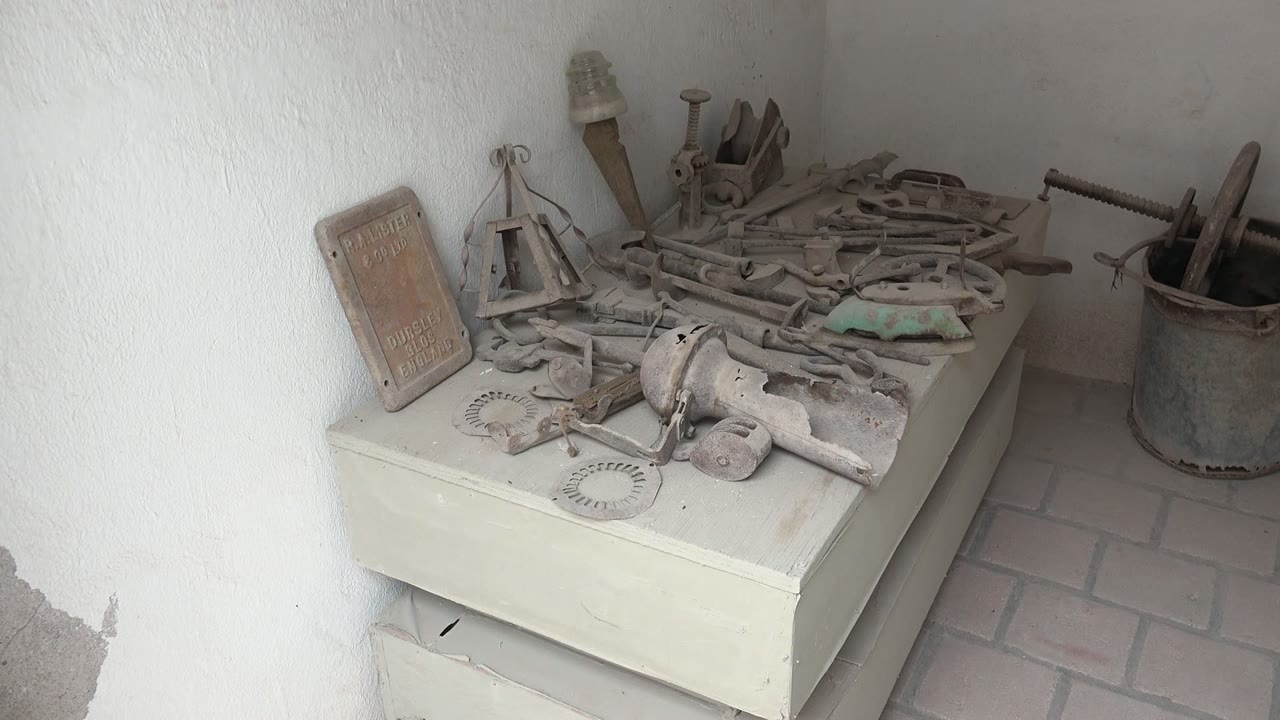 The cabinet remains stacked with dusty tools in the Mulege workshop, while the rusted bucket and long handles sit to the right against white plaster walls, with no visible people.