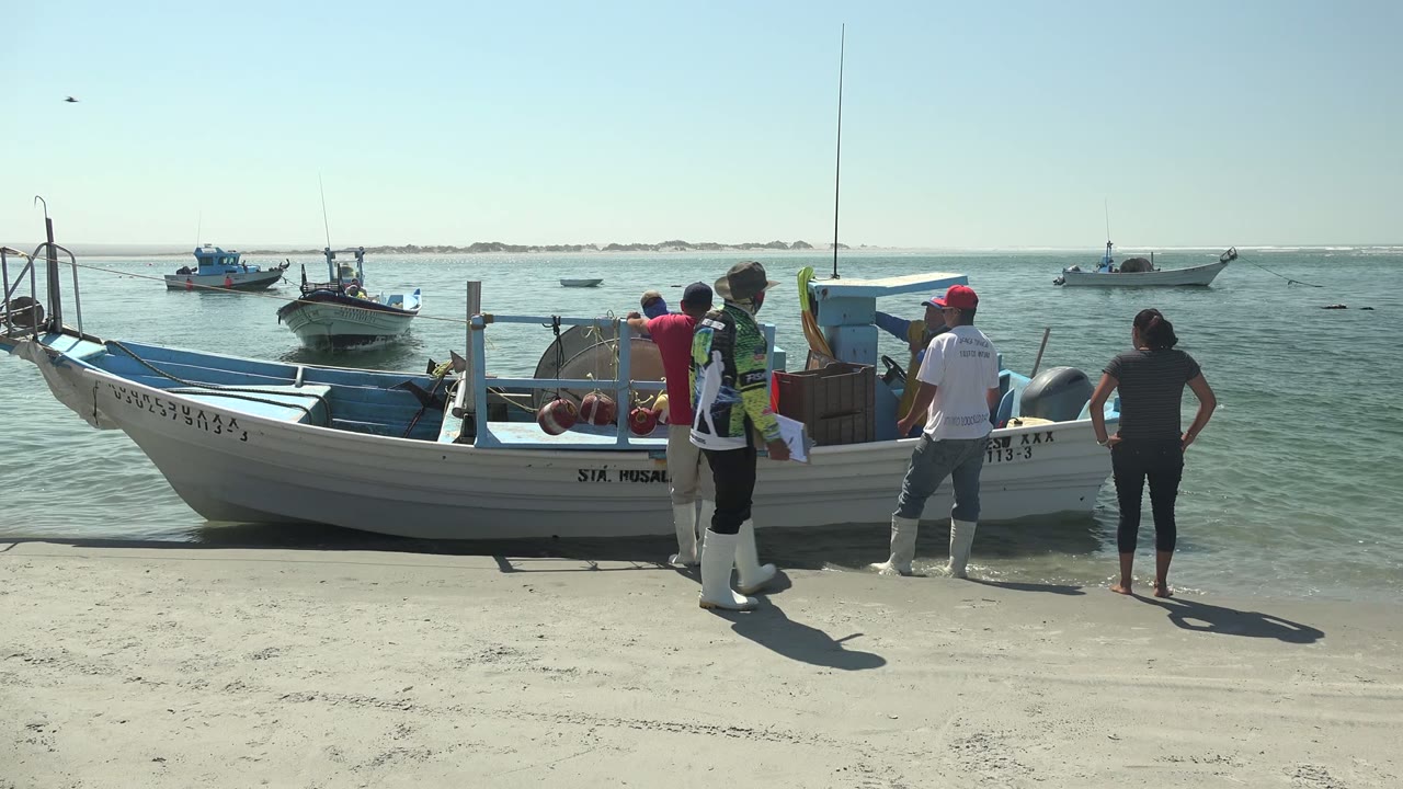 At La Bocana in Baja California Sur, several people stand beside a white and blue boat on pale sand while other boats sit on calm water offshore.