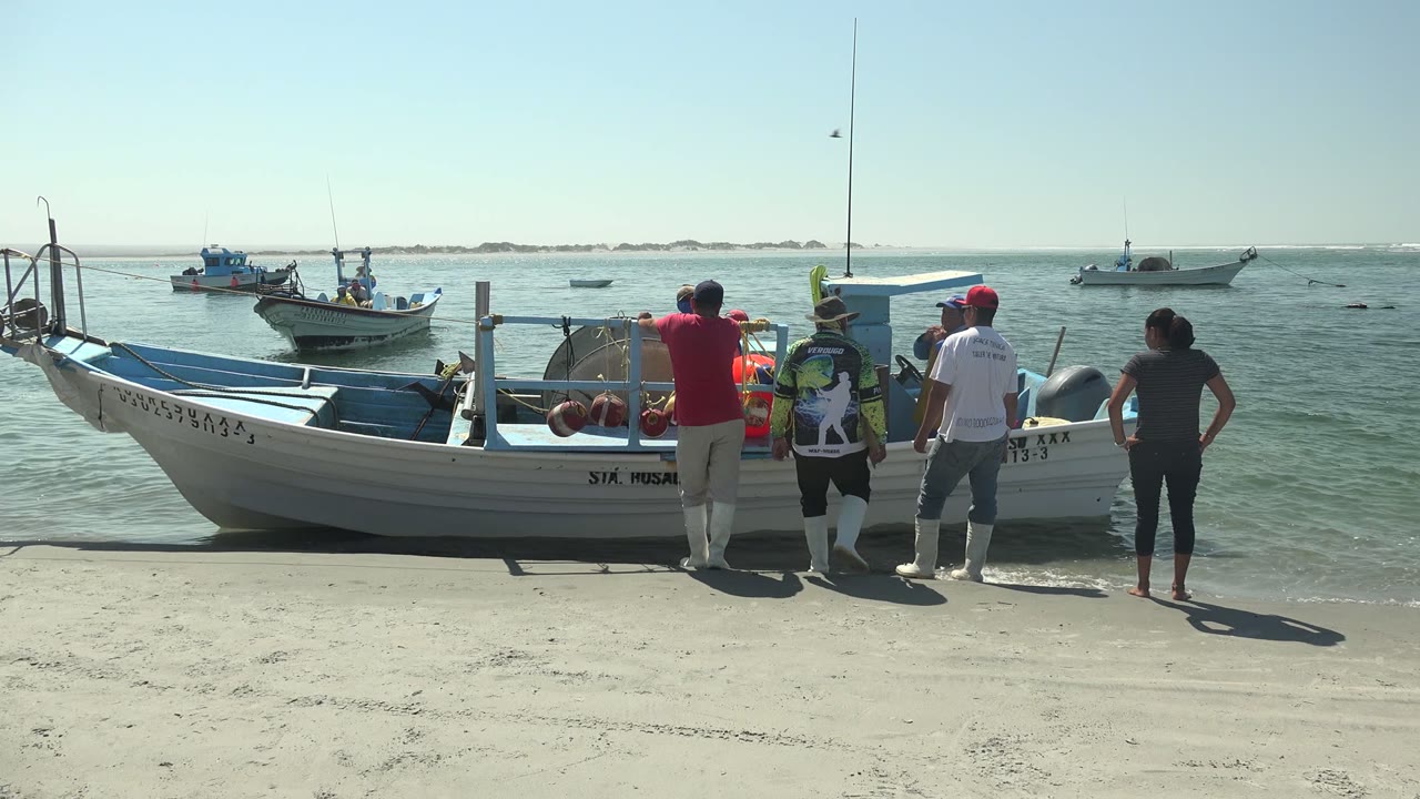 At La Bocana in Baja California Sur, the boat remains pulled up on sand while people stand along its beach side and small boats float across the calm water behind them.