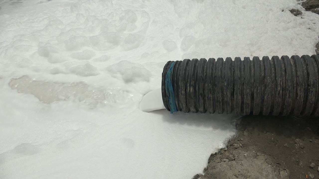 At 0:00 in Guerrero Negro, a black corrugated hose with a blue-edged opening releases white foam across pale ground, with no visible people.