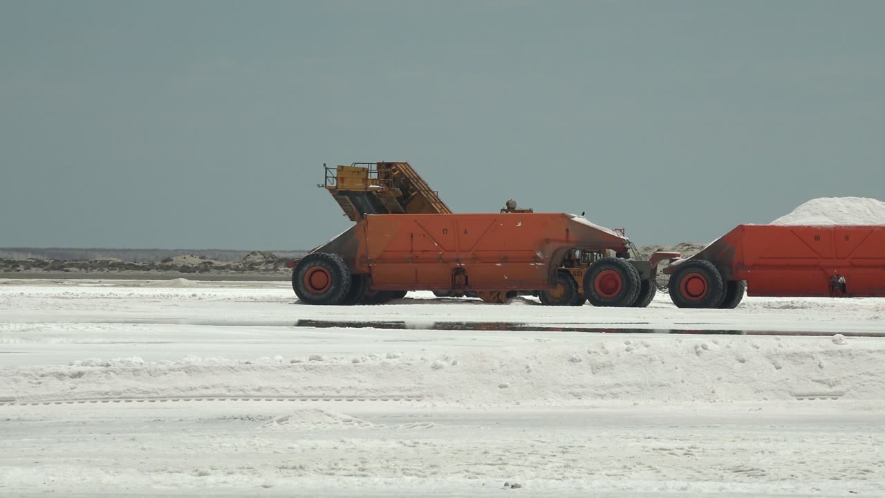 At the start of the clip in Guerrero Negro, a red industrial hauler pulls a red container loaded with white material across a flat white surface, with no visible people.